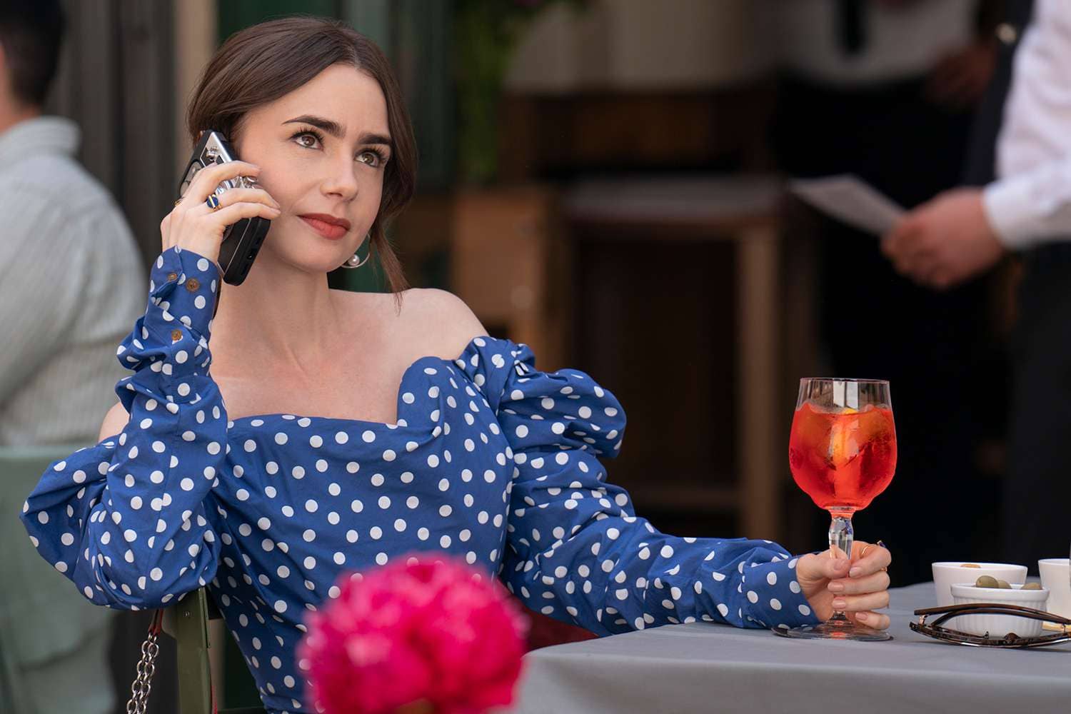 Emily in Paris, vêtue d'une robe à pois bleue, tient un verre de cocktail en main tout en parlant au téléphone dans un café parisien, montrant son style audacieux et son quotidien dans la ville lumière.