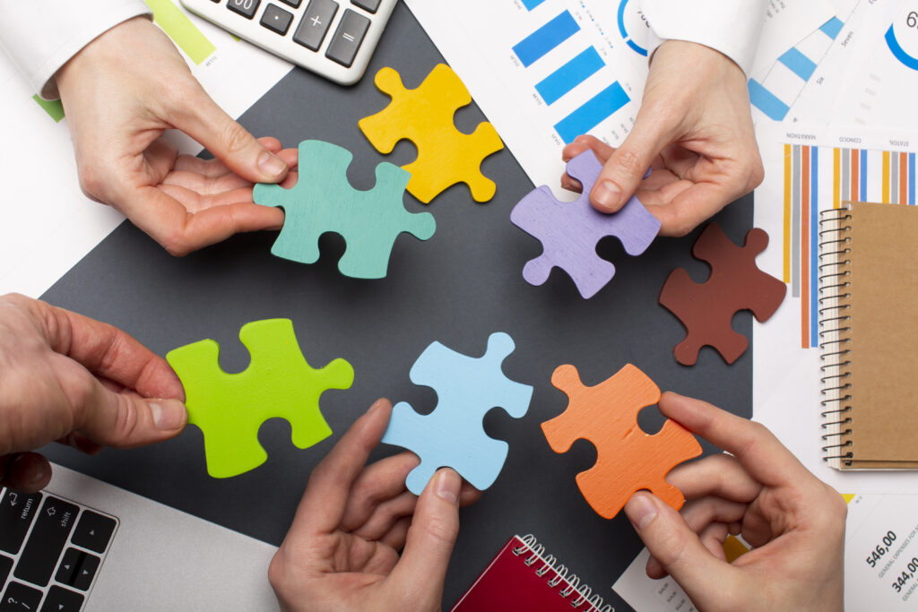 Cette vue de dessus montre plusieurs personnes réunies autour d'une table de travail. Chaque participant tient une pièce de puzzle en bois de couleur vive (vert, bleu, orange, violet, jaune) qu'ils rapprochent les unes des autres sur une surface sombre centralisée.

Autour d'eux se trouvent plusieurs éléments d'un environnement de bureau : des graphiques financiers, un ordinateur portable, une calculatrice et des carnets à spirales. L'ensemble évoque des concepts de collaboration, de travail d'équipe et de recherche de solutions en entreprise.